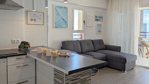 A modern kitchen with a grey sofa, a dining area, and a large window with sheer curtains.