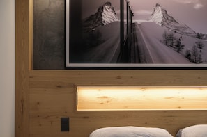 A framed photograph of a mountain road with snow-covered peaks, a wooden headboard, and a bedside lamp. - Hotel La Réserve (St. Niklaus)