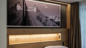 A hotel room with a large framed mountain landscape picture, a wooden headboard with built-in lighting, and a window with curtains. - Hotel La Réserve (St. Niklaus)