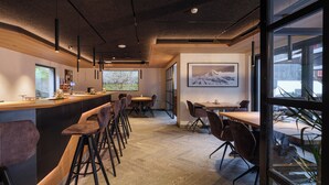 A modern bar area with a long counter, bar stools, and a large window offering a view of the outdoors. - Hotel La Réserve (St. Niklaus)