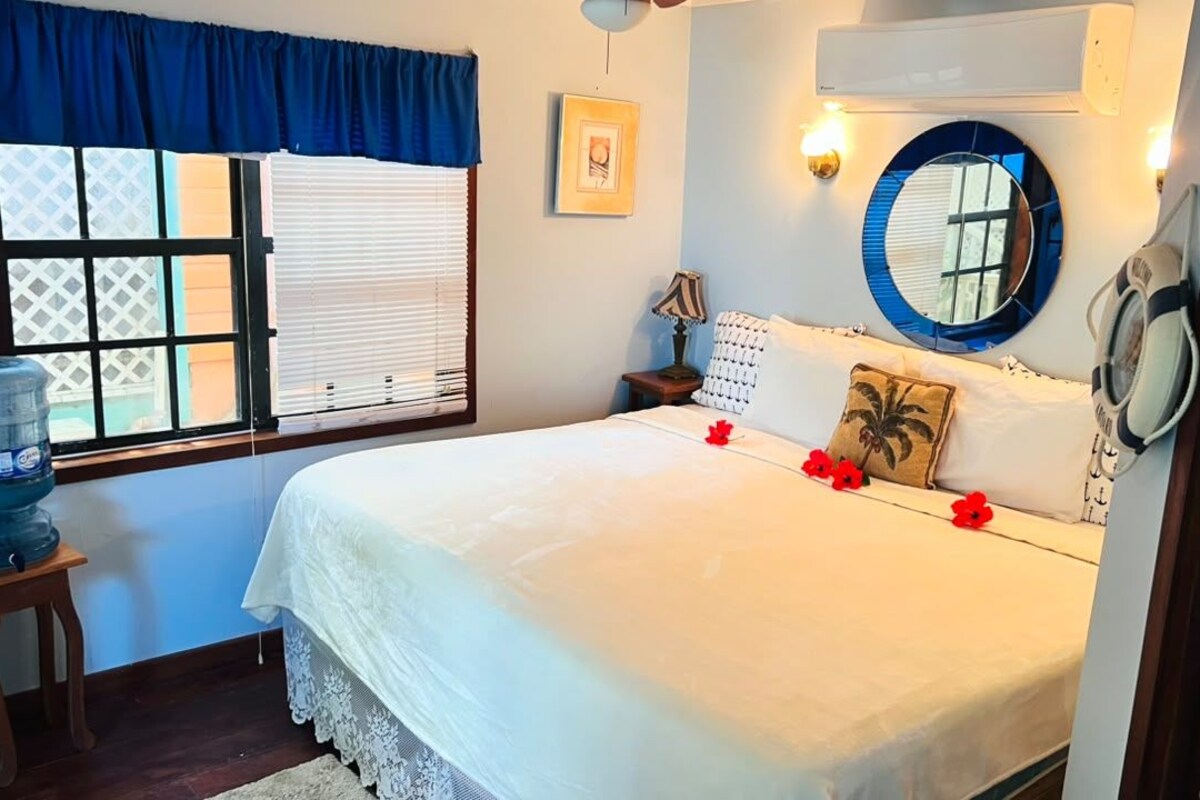 A bedroom with a bed, a water dispenser, a nightstand, a lamp, a mirror, and a window with blinds.