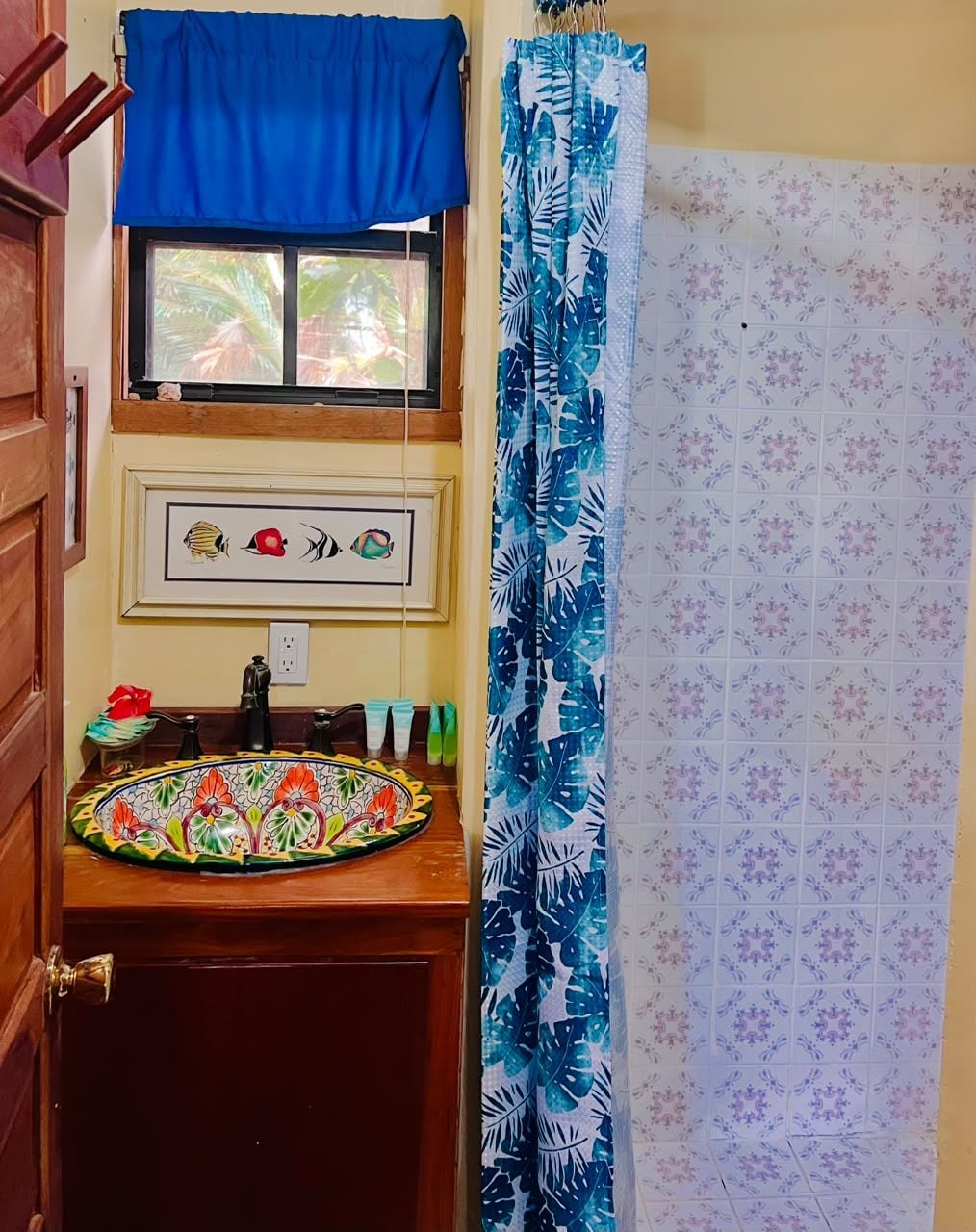 Un baño con tocador de madera, un lavamanos colorido, una ventana con cortina azul y una zona de ducha con cortina floral.