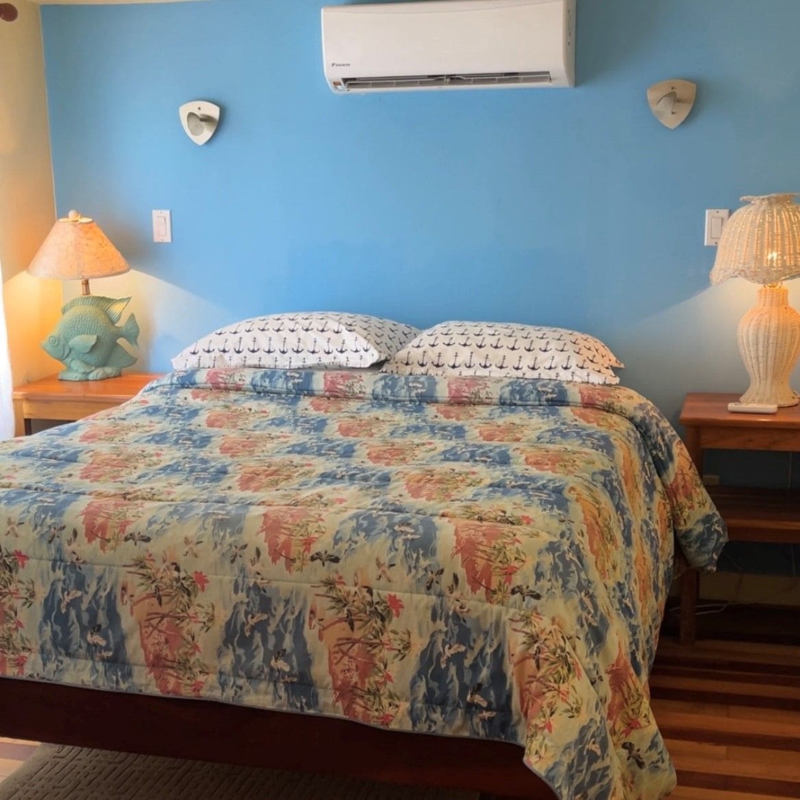 A bedroom with a floral bedspread, two decorative pillows, a wall-mounted air conditioner, and a small decorative fish on a nightstand.