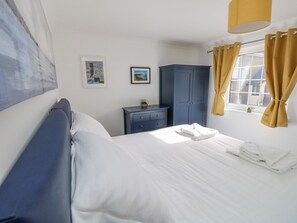 A neatly made bed with white linens, a dark blue wardrobe, and a small nightstand with a lamp. - Tennyson View (Totland Bay)