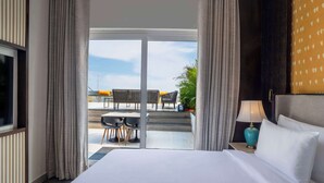 A hotel room with a large bed, a nightstand with a lamp, and a view of an outdoor seating area with chairs and a table.