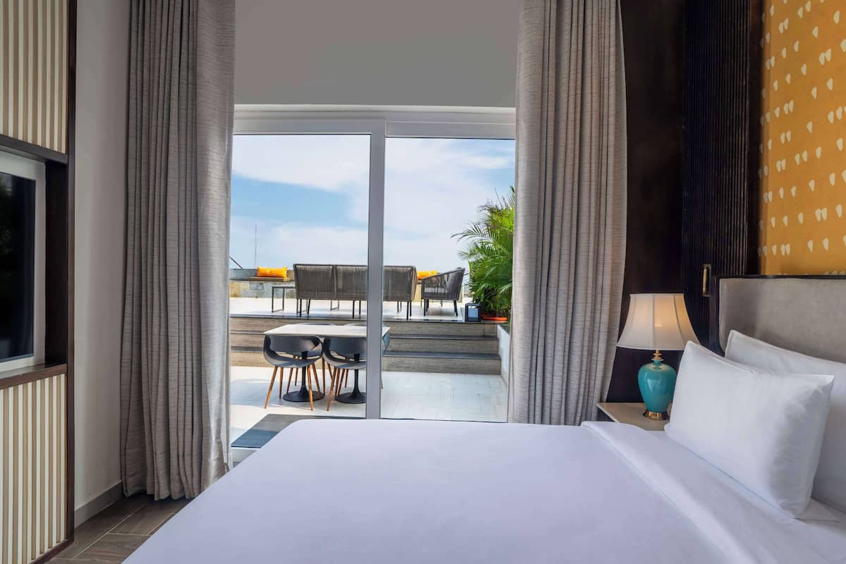A hotel room with a large bed, a nightstand with a lamp, and a view of an outdoor seating area with chairs and a table.