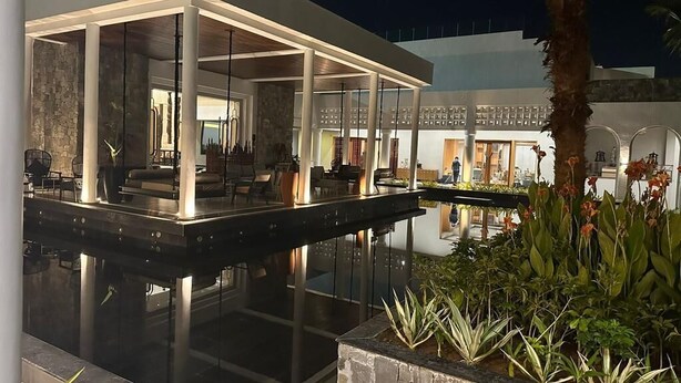 A modern outdoor pool area with a glass-enclosed lounge, surrounded by plants and a stone structure.