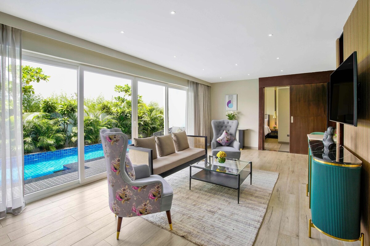 A modern living room with a sofa, armchair, and coffee table, large windows with a view of a swimming pool and greenery, and a flat-screen TV mounted on the wall.