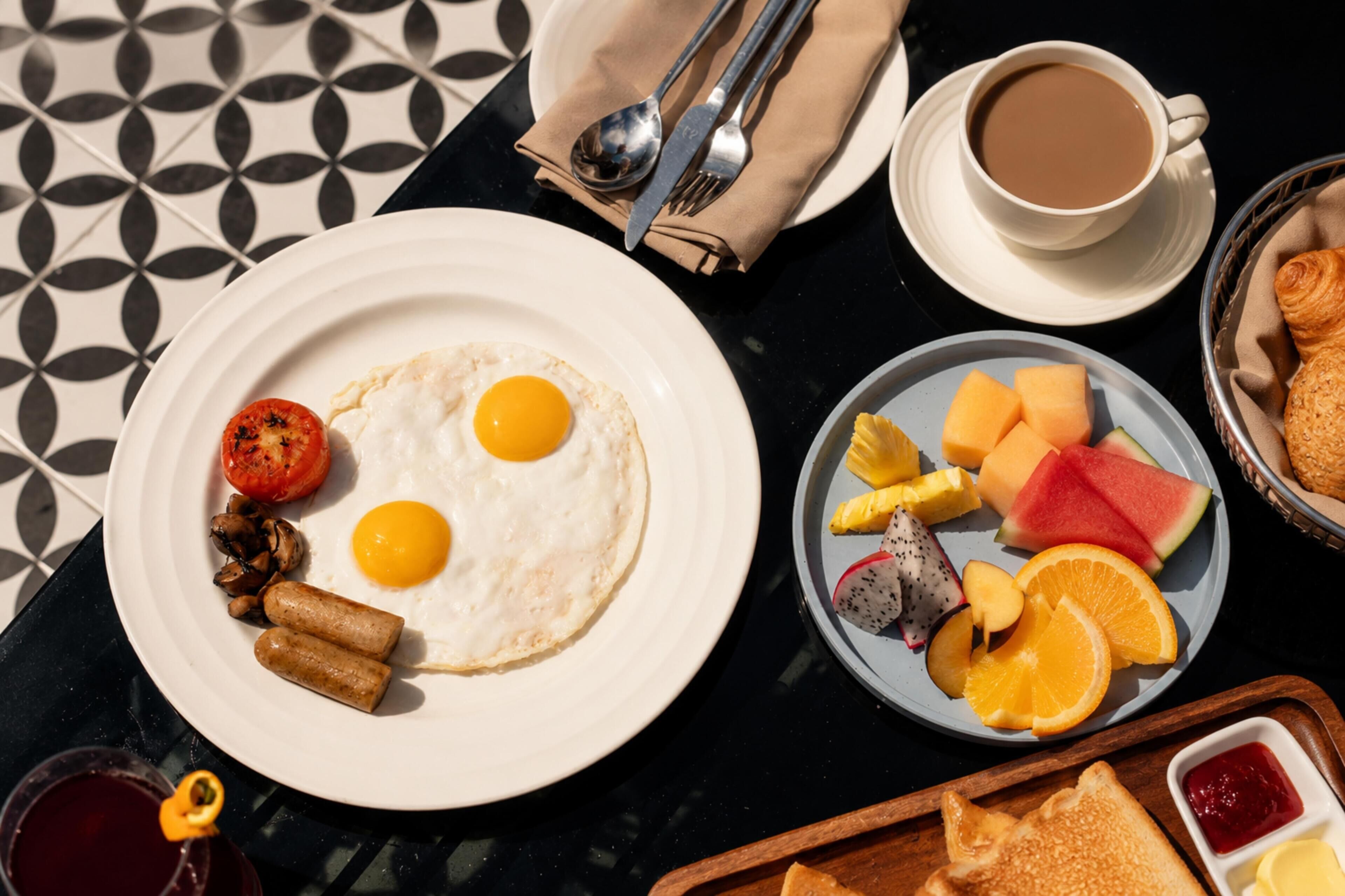Un desayuno con huevos, salchichas y tostadas en un plato, un tazón de fruta, una taza de café y un vaso de bebida roja.