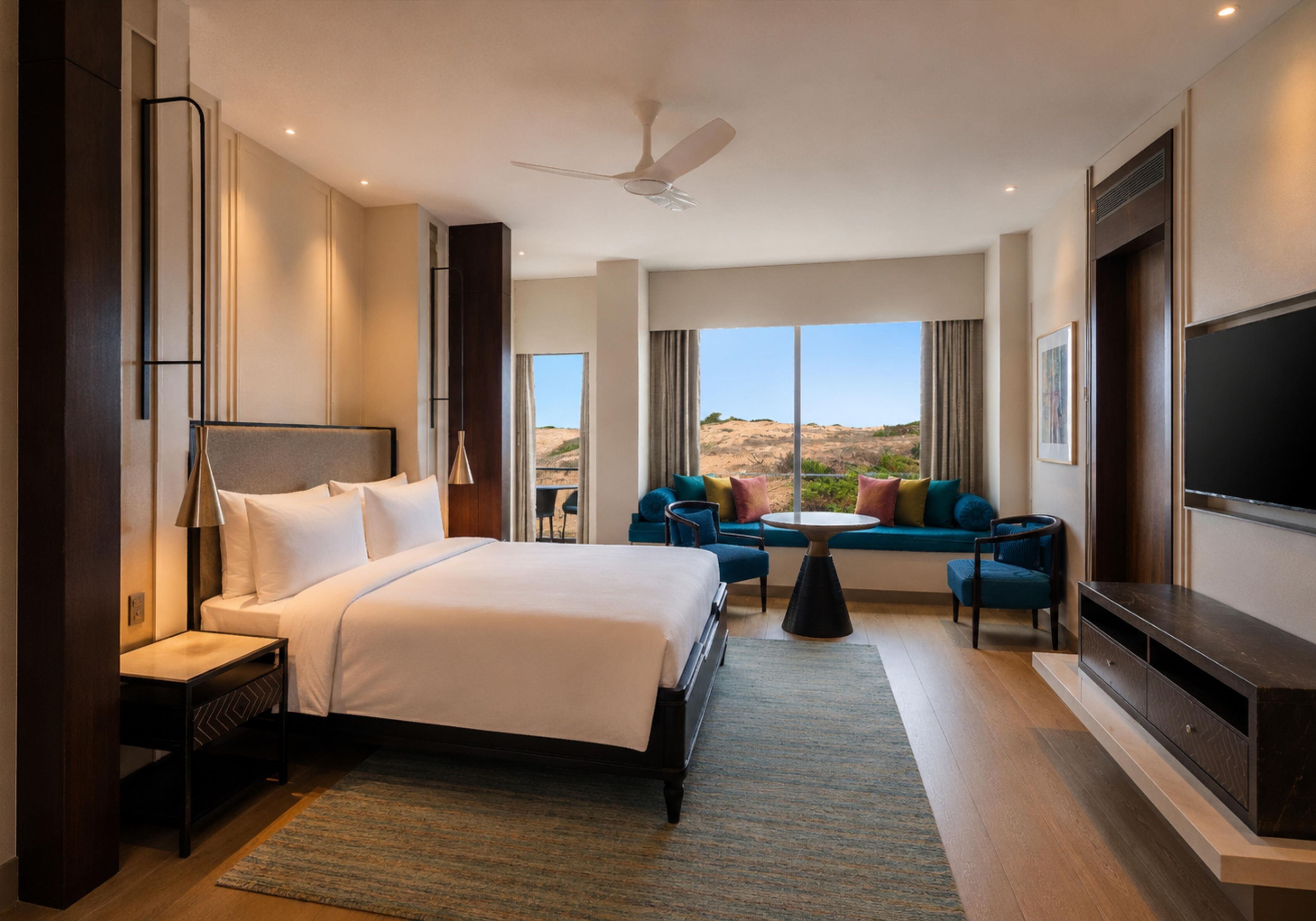 A modern hotel room with a large bed, a seating area with a sofa and chairs, a flat-screen TV, and a view of the outdoors.