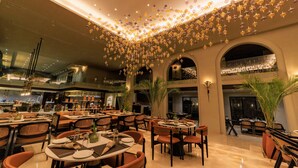 A spacious dining area with multiple tables and chairs, a high ceiling with hanging decorations, and a view of the kitchen in the background.