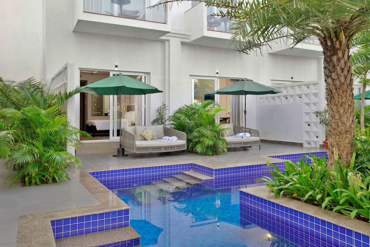 A pool area with blue-tiled steps, two outdoor sofas with green umbrellas, and a palm tree.