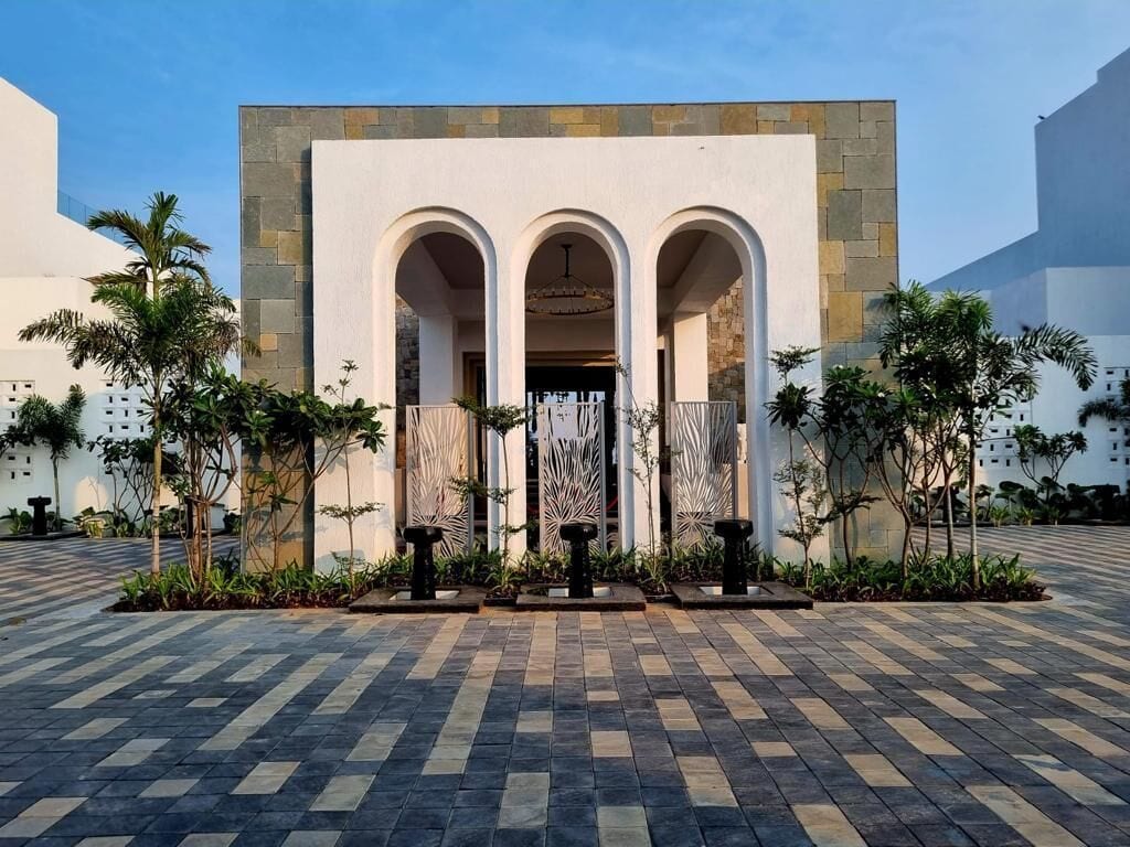 A building with a central arched entrance, flanked by columns, and a tiled courtyard with palm trees.