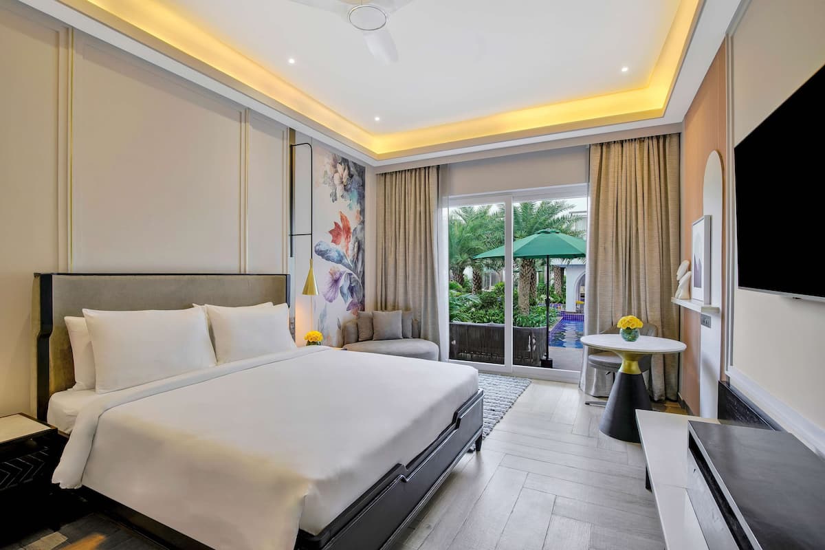 A hotel room with a large bed, a TV, a small table with a vase, and a view of the outdoors.