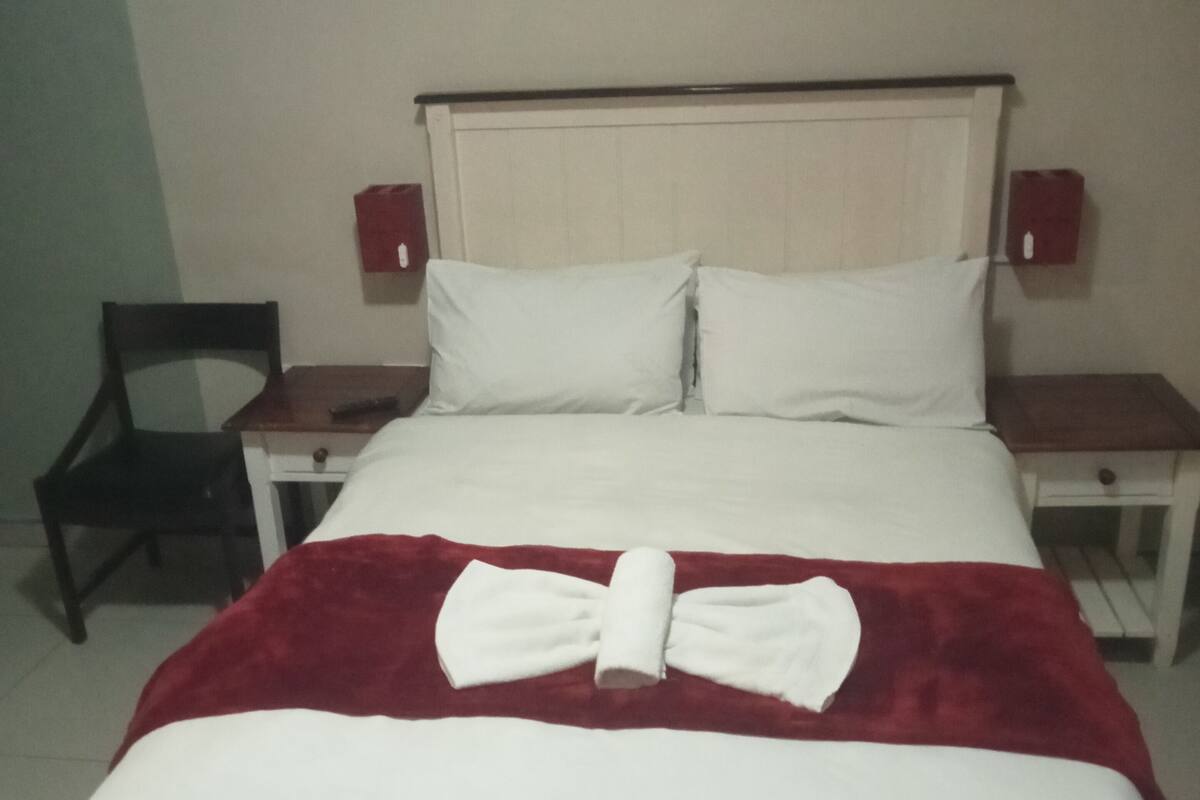 Een net opgemaakt bed met een wit-rode deken, twee witte kussens en een handdoek gevouwen in een strik. Aan beide zijden van het bed staan nachtkastjes met lampen.