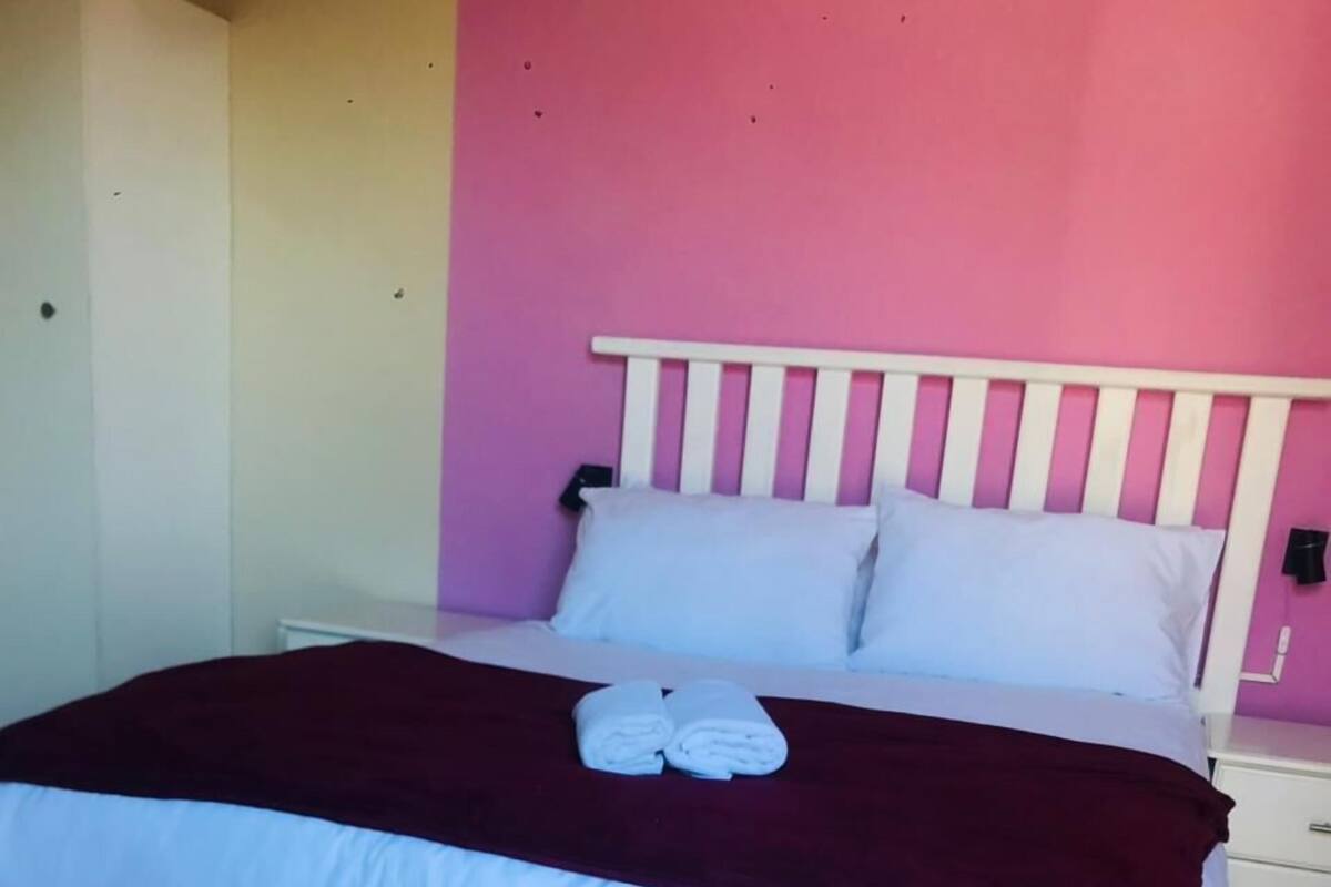 Een bed met een wit hoofdeinde, een rode deken en twee opgerolde handdoeken.