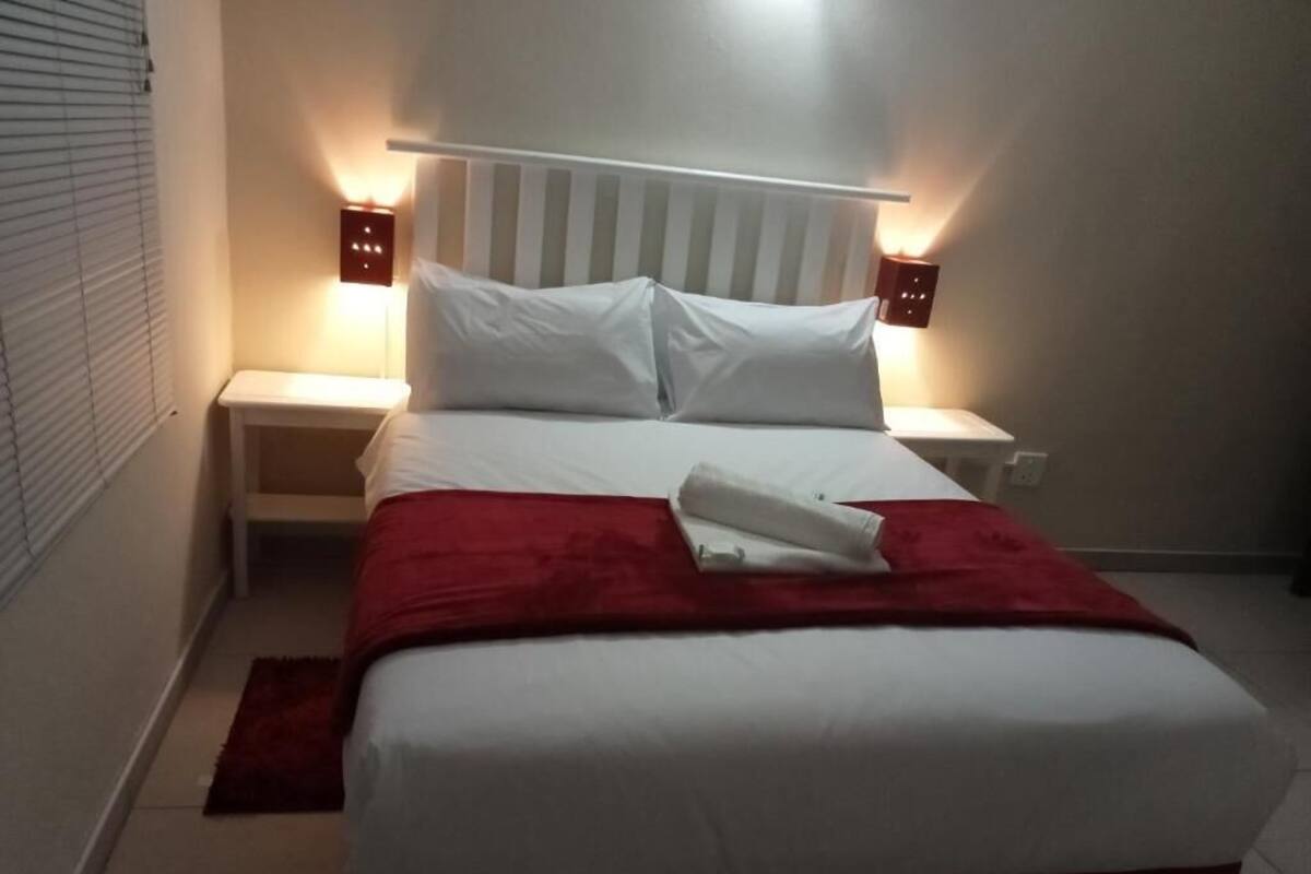 Een net opgemaakt bed met wit en rood beddengoed, twee nachtkastjes met lampen en een rood vloerkleed.