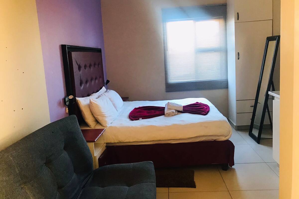 Een slaapkamer met een bed, een stoel, een bureau, een raam met jaloezieën en een kledingkast.