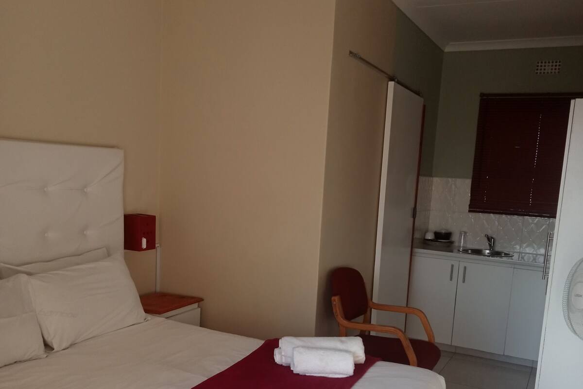 Een hotelkamer met een bed, een rode deken, een stoel, een klein tafeltje met een lamp, en een kitchenette op de achtergrond.