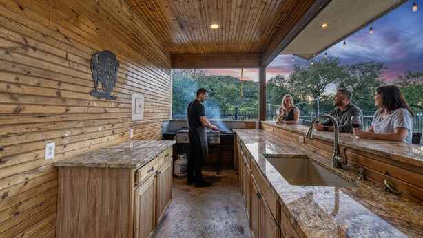 A modern kitchen with wooden walls, a granite countertop, and a large window overlooking a scenic landscape. - Tatanka @ Frio Premier Rental Homes (Concan)
