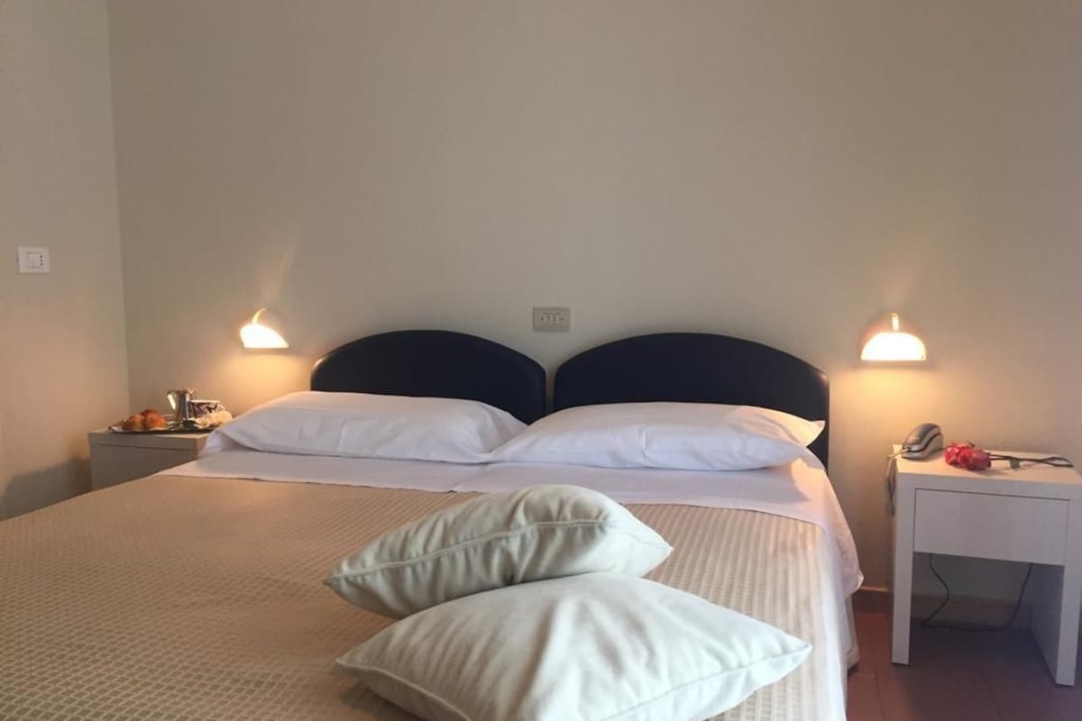 A neatly made bed with white linens and two pillows, a bedside table with a lamp, and a small desk with a telephone and a red object.