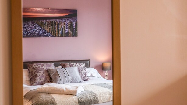 A bedroom with a bed, pillows, a blanket, a nightstand, a lamp, and a painting of a landscape on the wall. - Chandler's View (Whitby)
