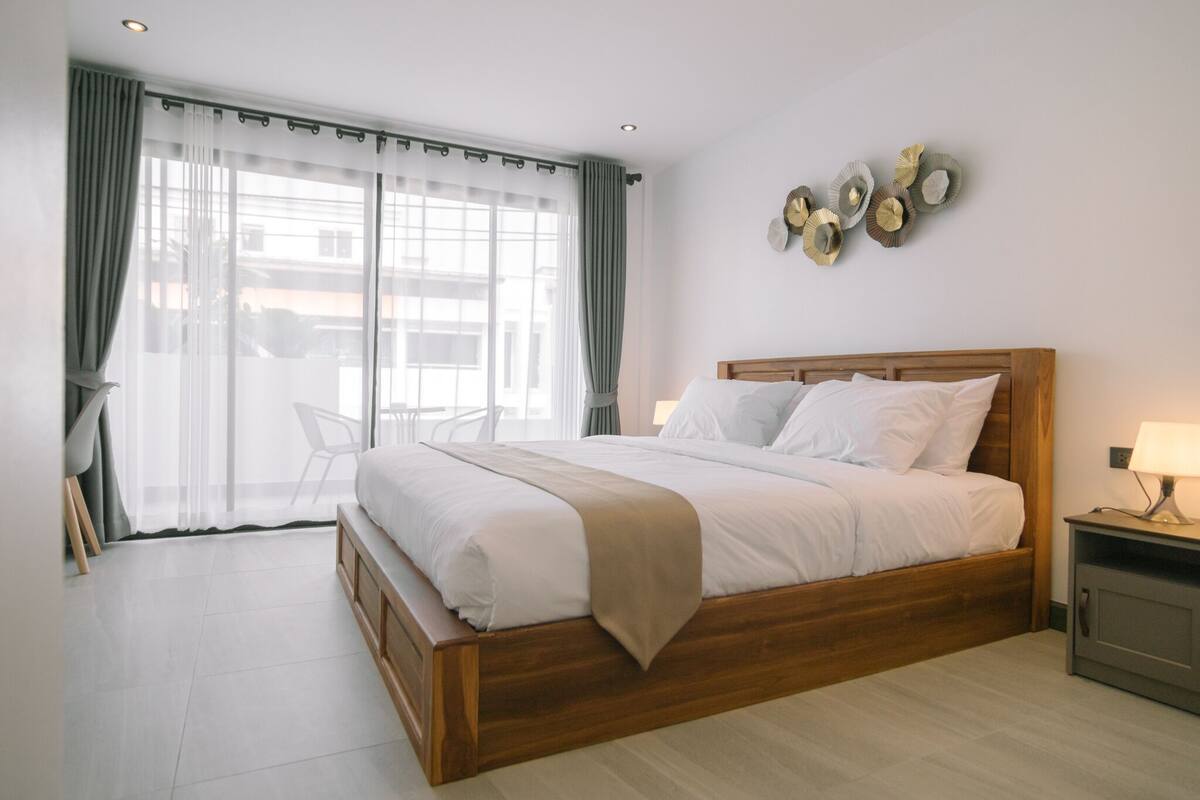 A neatly arranged bedroom with a wooden bed, white bedding, a bedside table with a lamp, and a view through sheer curtains.