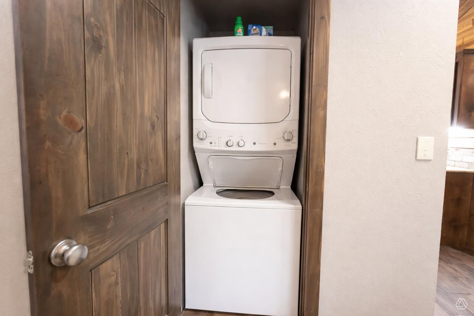 A compact laundry room with a washer and dryer, wooden door, and a small shelf with cleaning supplies.