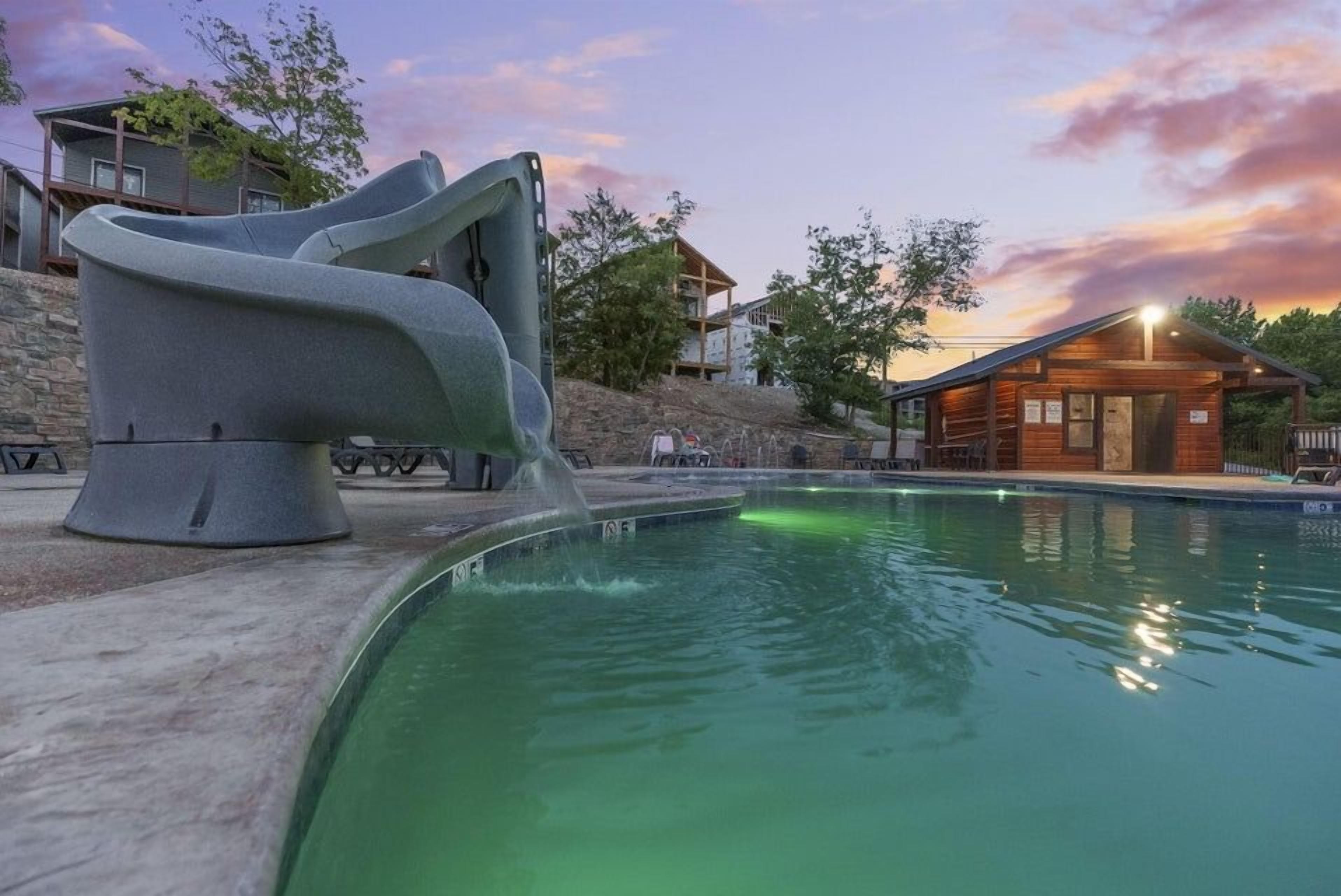 A swimming pool with a unique slide structure, surrounded by buildings and a sunset sky.