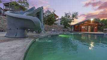 A swimming pool with a unique slide structure, surrounded by buildings and a sunset sky.