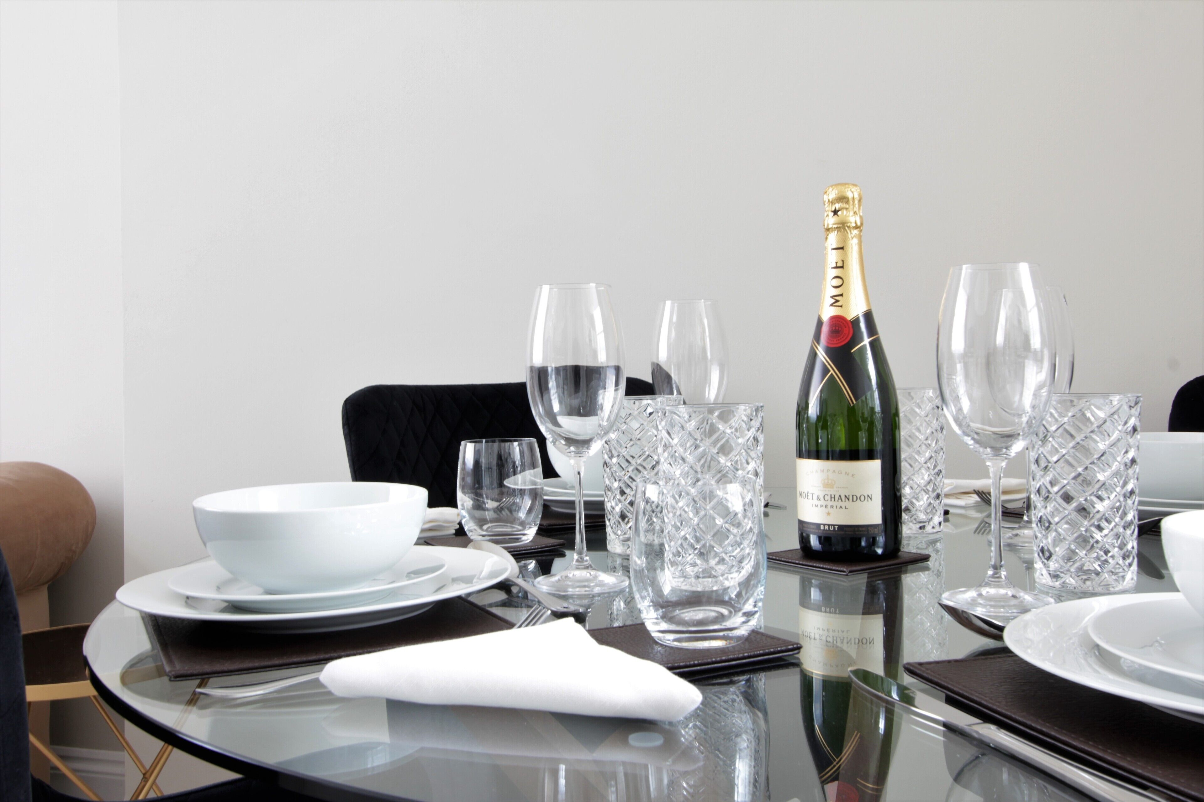 Une table à manger dressée avec de l’assiette blanche, des bols et de la verrerie transparente, ainsi qu’une bouteille de champagne.