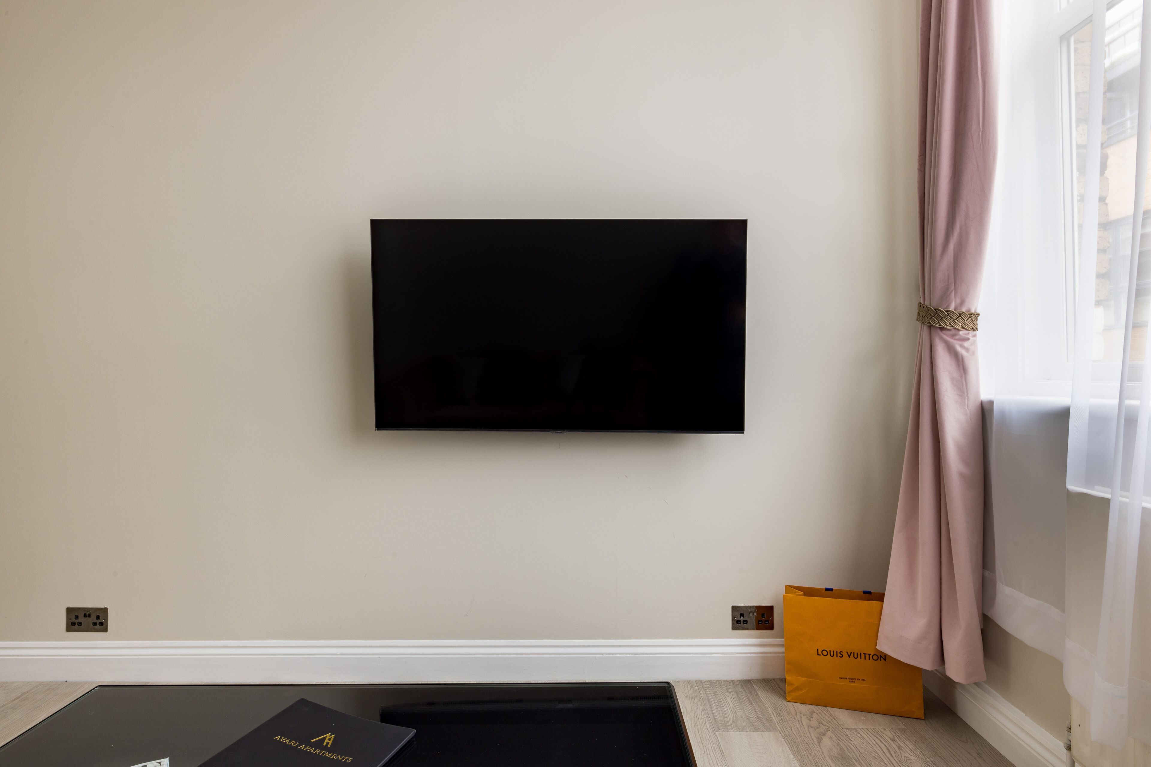 A flat-screen TV mounted on a wall, a black box labeled 'AVARI APARTMENTS,' and a Louis Vuitton shopping bag on the floor.