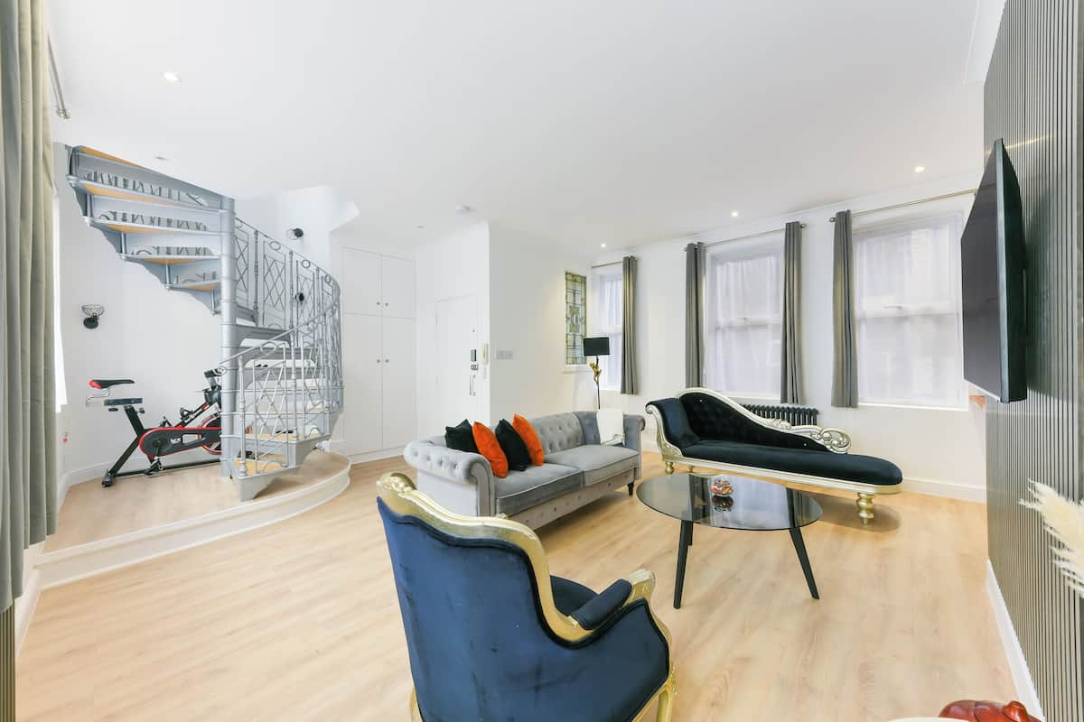 A modern living room with a staircase, a grey sofa, a blue armchair, and a dining table.