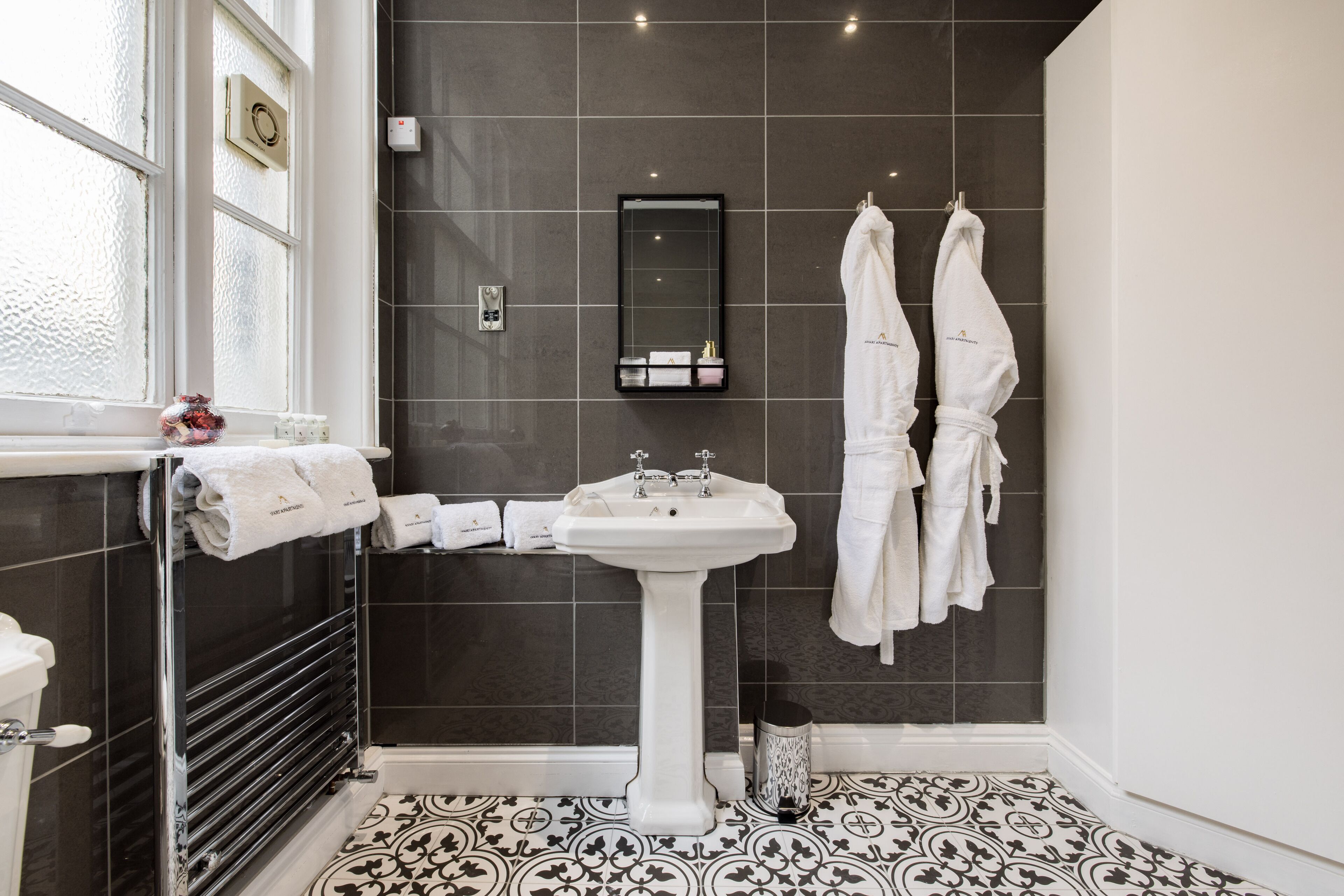 Une salle de bain moderne avec un lavabo sur colonne blanc, des murs recouverts de carreaux foncés et un sol à motifs.