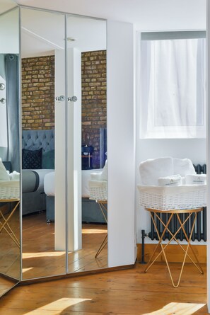 Une chambre d’hôtel moderne avec un placard aux portes miroir, un mur en briques, un lit doté d’une tête de lit capitonnée, une table de chevet en osier blanc et un radiateur.
