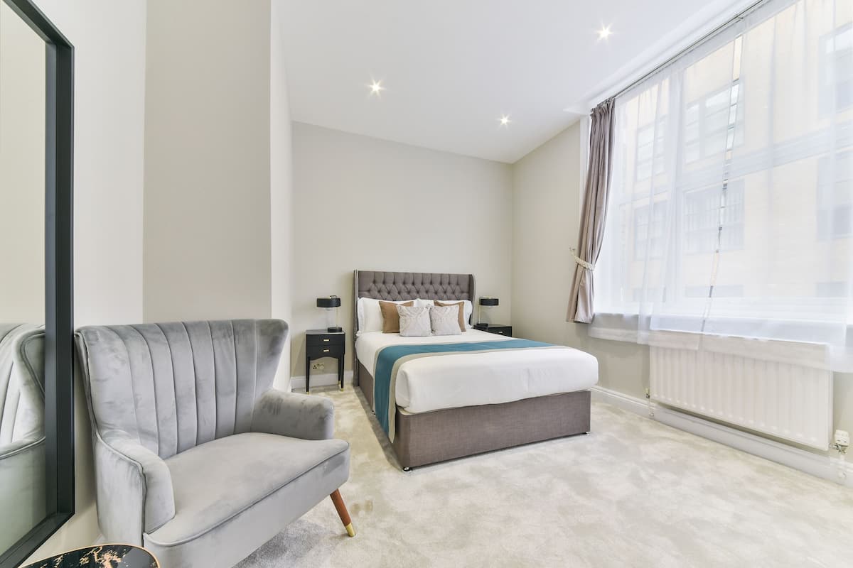 A modern bedroom with a grey sofa, a large bed with a grey headboard, bedside tables with lamps, and a large window with sheer curtains.