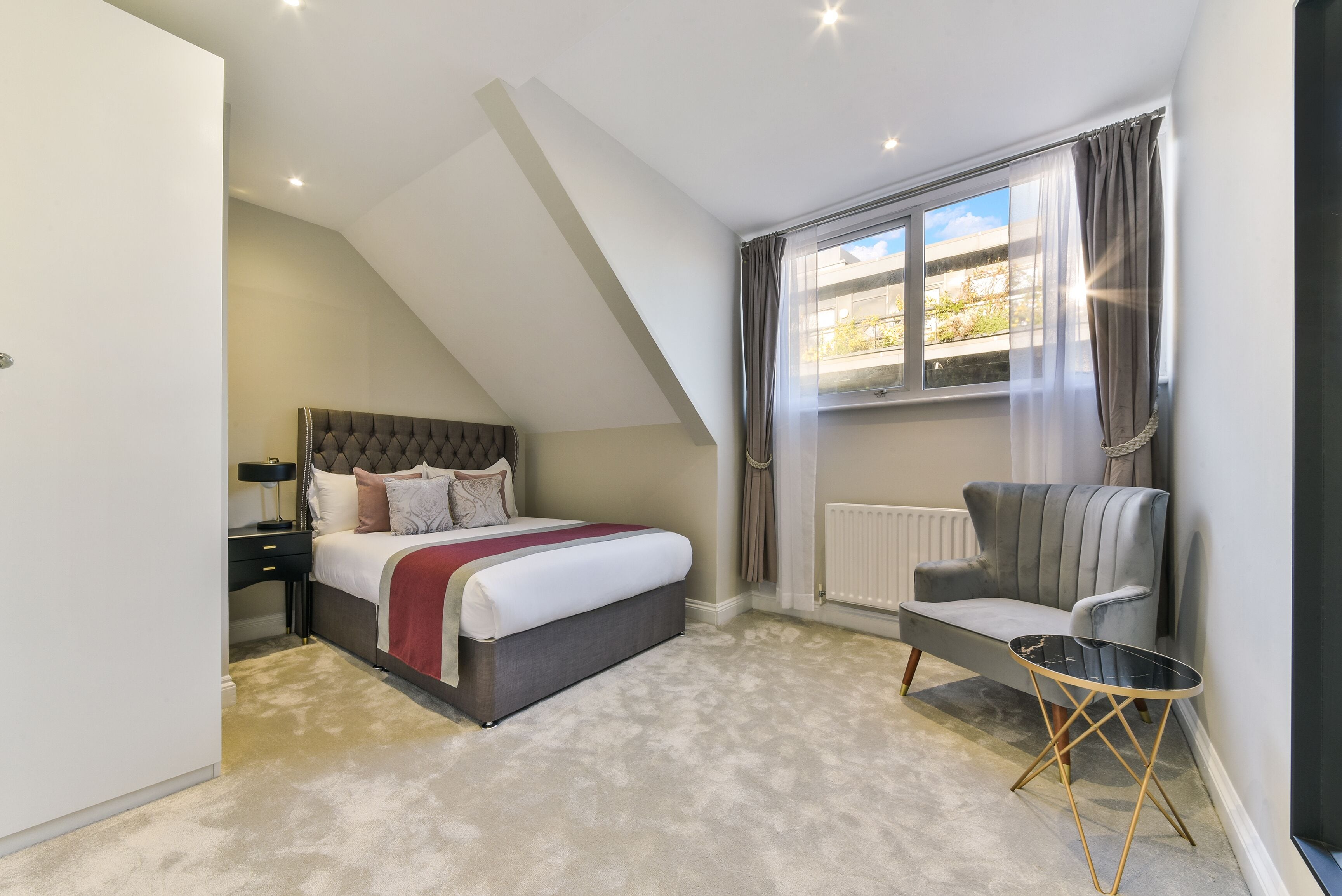 A bedroom with a bed, a chair, a small table, and a window with curtains.