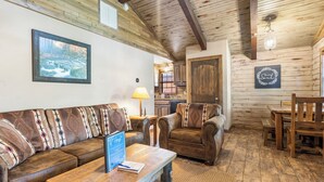 Una habitación acogedora con techo de madera, un sofá con cojines estampados, una mesa de centro de madera con un cartel, y un área de comedor con sillas y mesa de madera.