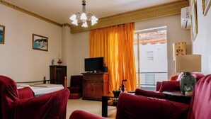 Een woonkamer met een televisie, rode meubels, een houten kast en een groot raam met oranje gordijnen.