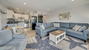 A modern living room with a grey sectional sofa, a coffee table, and a dining area with a white table and chairs. A kitchen with white cabinets and appliances is visible in the background. - Newport Bay 5 by Seaside Vacations & Sales (Ocean City)