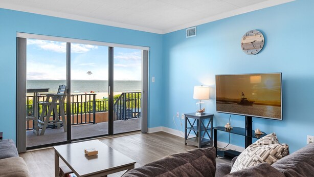 A living room with a view of the ocean, a clock on the wall, and a TV on a stand.