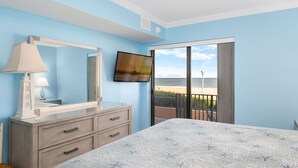 A bedroom with a bed, a dresser, a mirror, a TV, and a balcony with a beach view.