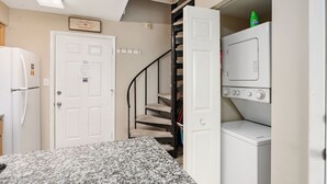 Uma lavanderia com máquina de lavar e secadora, uma porta branca com um aviso, uma escada com um arranhador para gatos e uma geladeira.