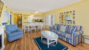Un salon avec un fauteuil bleu, un canapé à rayures, une table basse en verre et un coin repas avec des chaises blanches, et une cuisine en arrière-plan.