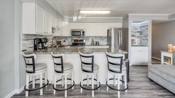 A modern kitchen with white cabinets, a granite countertop, a stainless steel refrigerator, and a microwave. There are three bar stools with black metal frames and white seats.