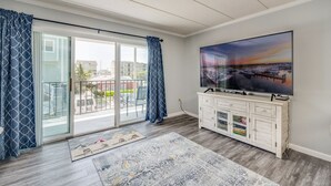 A spacious living room with a large TV, a sliding glass door leading to a balcony, and a view of the cityscape.