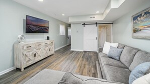 A spacious living room with a large flat-screen TV, a wooden cabinet, a sliding barn-style door, and a gray sofa with patterned cushions. - Siesta Villa 101N by Seaside Vacations & Sales (Ocean City)