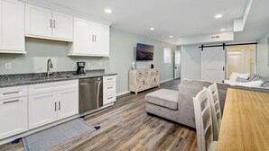 A modern kitchen with white cabinets, a stainless steel dishwasher, and a granite countertop. There is a dining area with a wooden table and chairs, and a gray sofa. - Siesta Villa 101N by Seaside Vacations & Sales (Ocean City)