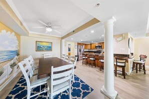 A dining area with a wooden table and white chairs, a kitchen with wooden cabinets, and a ceiling fan. - Somerset on 2nd 202 by Seaside Vacations & Sales (Ocean City)