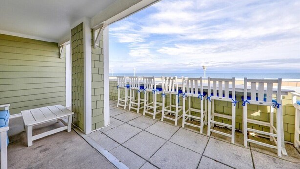 Une terrasse couverte avec vue sur la plage, des chaises blanches ornées de rubans bleus et une petite table.